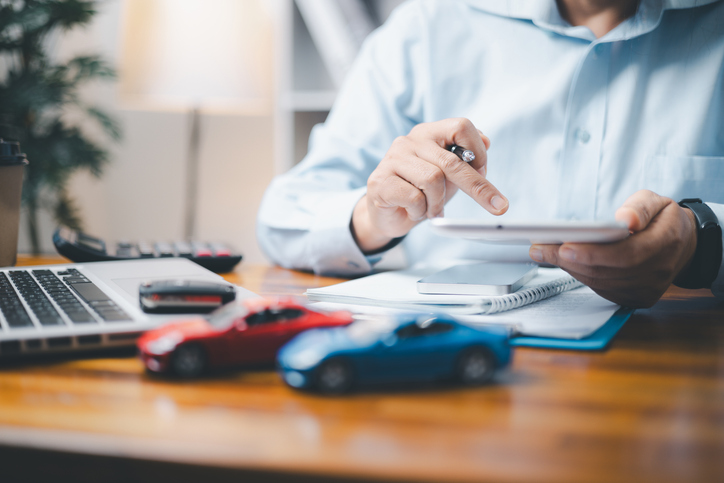 Businessman sits at a desk with a laptop, clipboard and two toy cars. He's inputting numbers into a calculator.