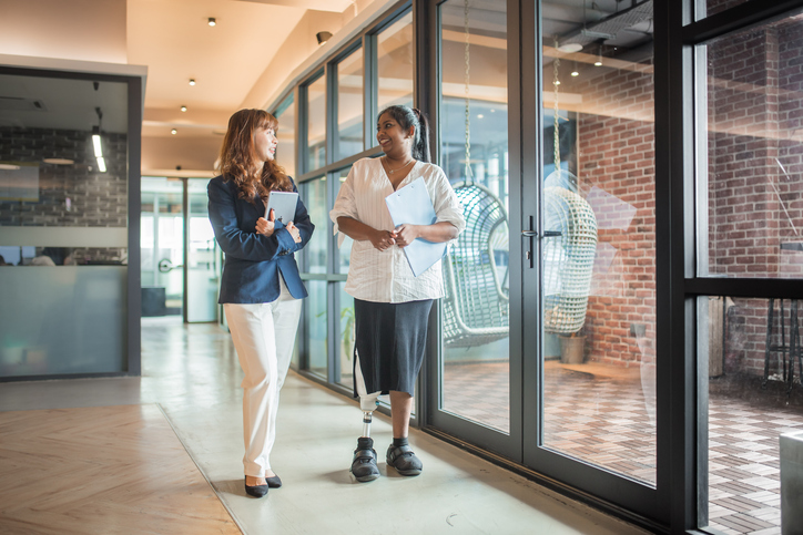 Asia Indian female handicapped businesswomen with prosthetic limb holding document and walking at the office corridor. She is discussing with her colleague alternate text for this image