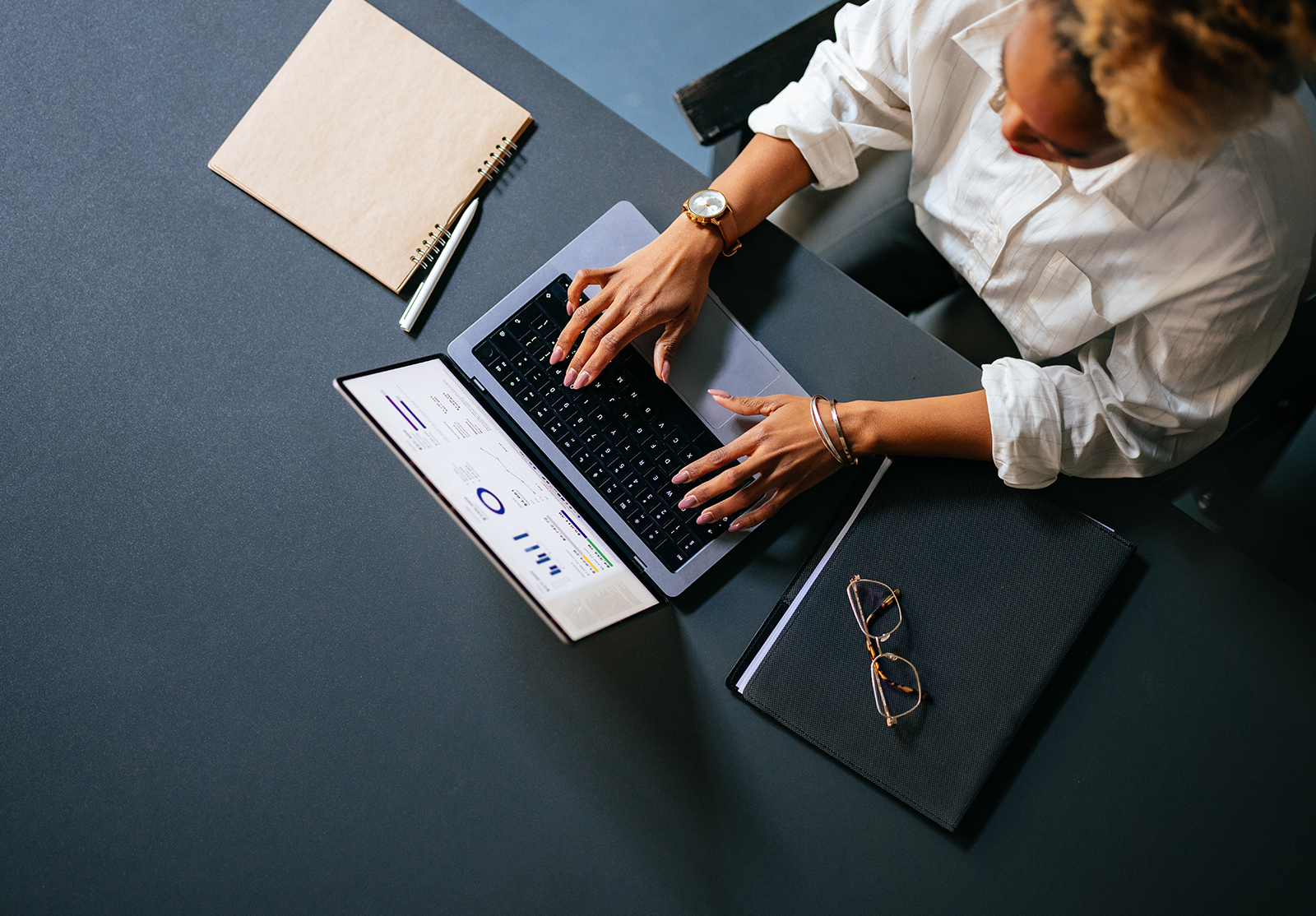 Person typing on a laptop with charts on screen, desk items include notebook, pen, glasses.