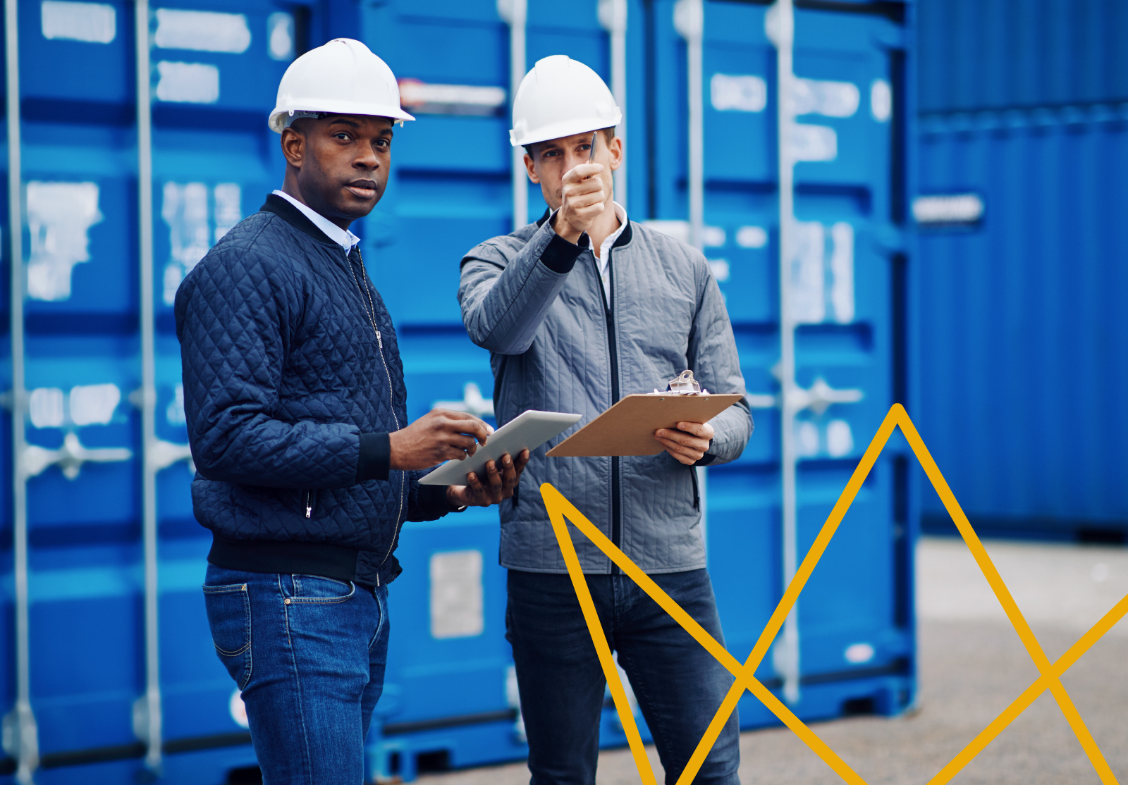 Two people in hard hats stand in front of stacked blue shipping containers, one with a tablet, the other pointing with a clipboard.