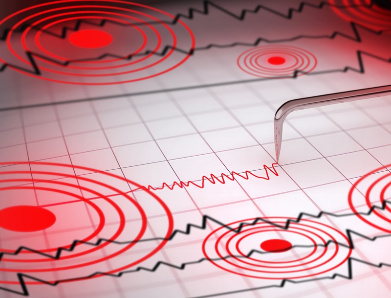 Close-up of a seismograph machine needle tracing seismic waves, indicative of earthquake activity, on graph paper with epicenter rings.