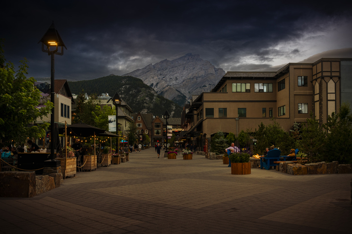 Banff, Canada - August 12, 2023: People walking along a street with shops and Restaurants. Some sitting around a campfire. In background majestic mountains.