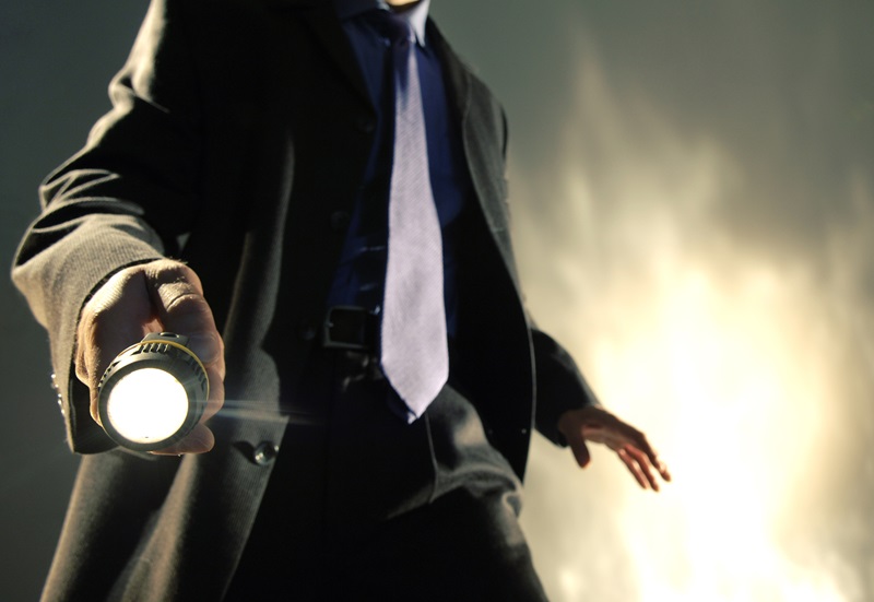 A close up of the torso of a Caucasian man dressed in a dark business suit, shirt and tie holding a battery operated torch pointed towards the camera, with his arm out in a dramatic pose. The man is standing against dark background with a brightly lit abstract pattern.
