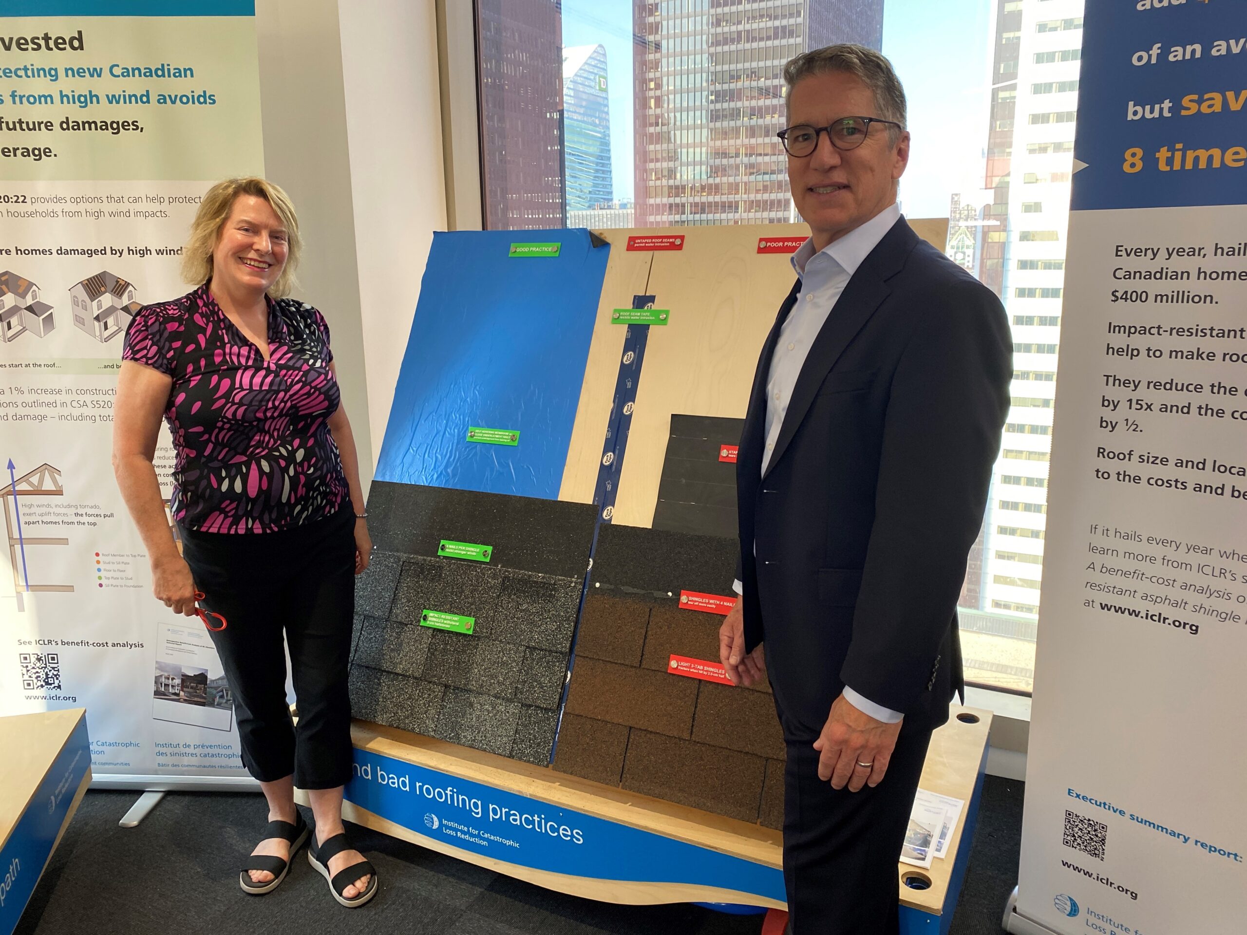 Phil Gibson, Senior Executive Advisor, Accenture Canada, and Moira Gill, Director of Strategy, ICLR, pose in front of a roof shingle display at the ICLR's resilience display centre in Accenture's Toronto office
