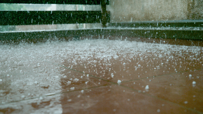 CLOSE UP: Tiny grains of soft hail fall on the wet brown floor of a balcony alternate text for this image
