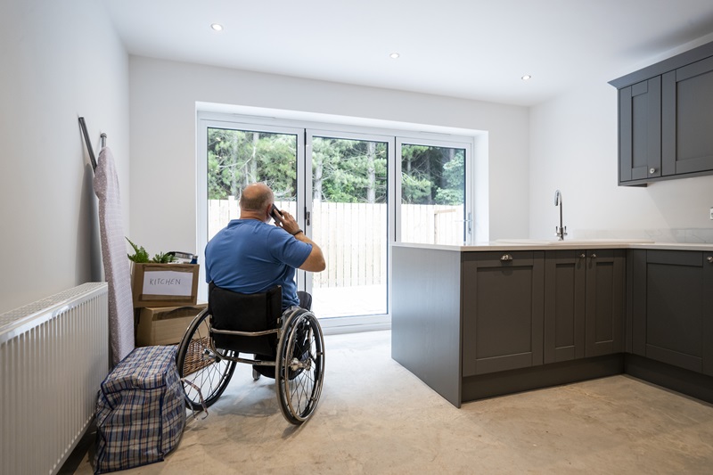 A rear-view shot of a mature man taking a break from unpacking cardboard boxes filled with his belongings in the house he has just bought. The house is incomplete and ready for renovation by the buyer. The man is a wheelchair user and he's in the kitchen.