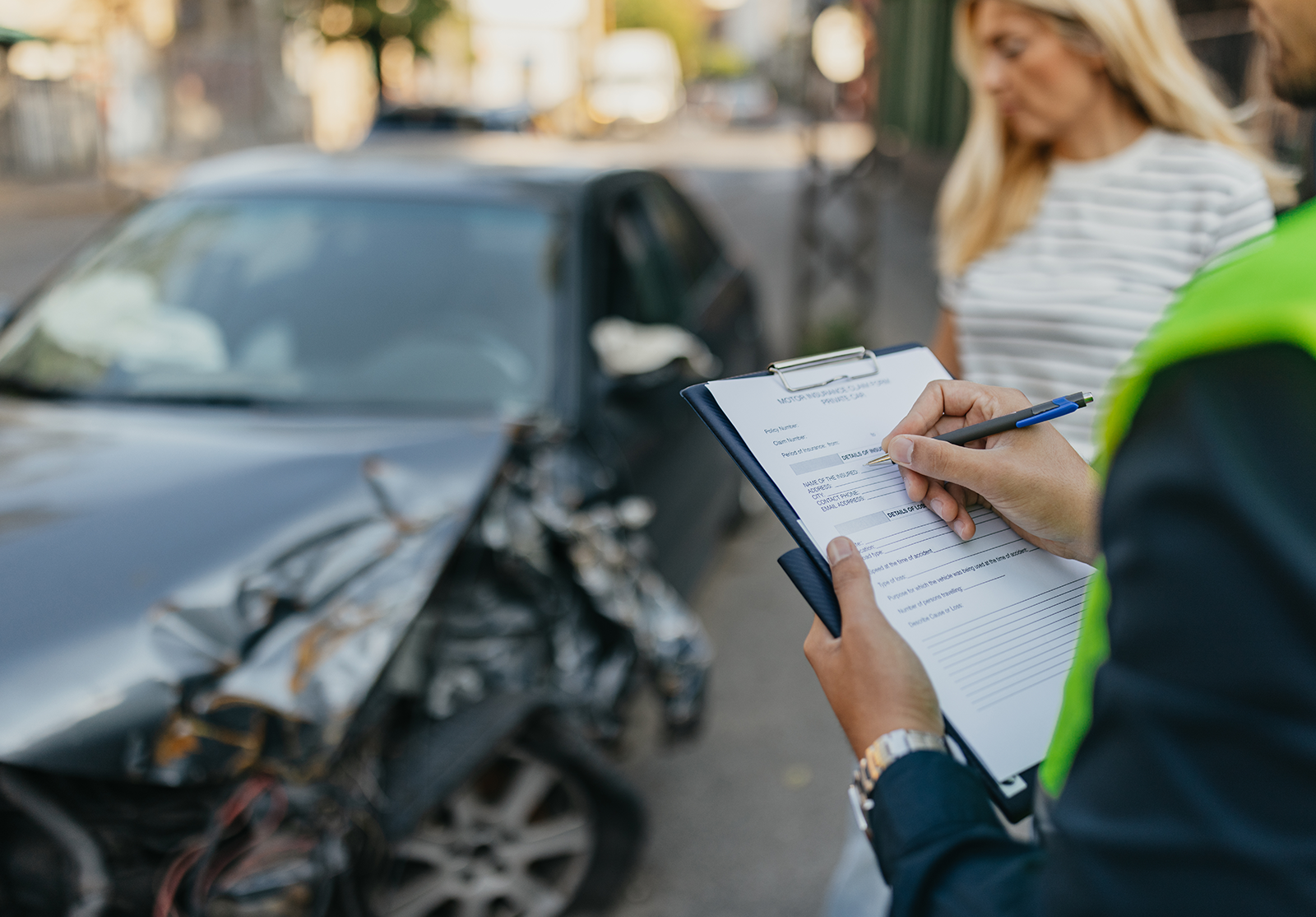 A person in a high-visibility vest documents a car accident beside a black vehicle with severe front-end damage on an urban street.