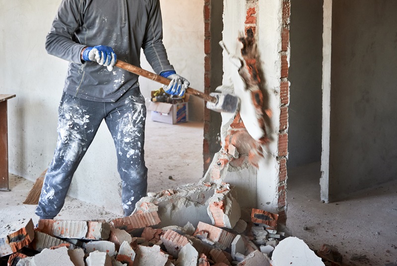 Close up of sledgehammer blow on brick, plaster. Workman striking devastating blow at remnants of interoom wall against backdrop of table and other plastered walls.