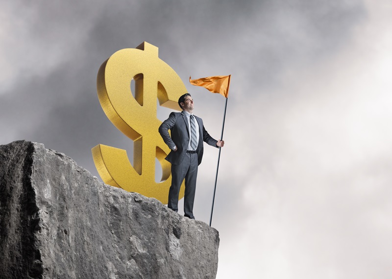 A man plants his flag on top of a tall cliff as he stands in front of a large gold dollar sign.