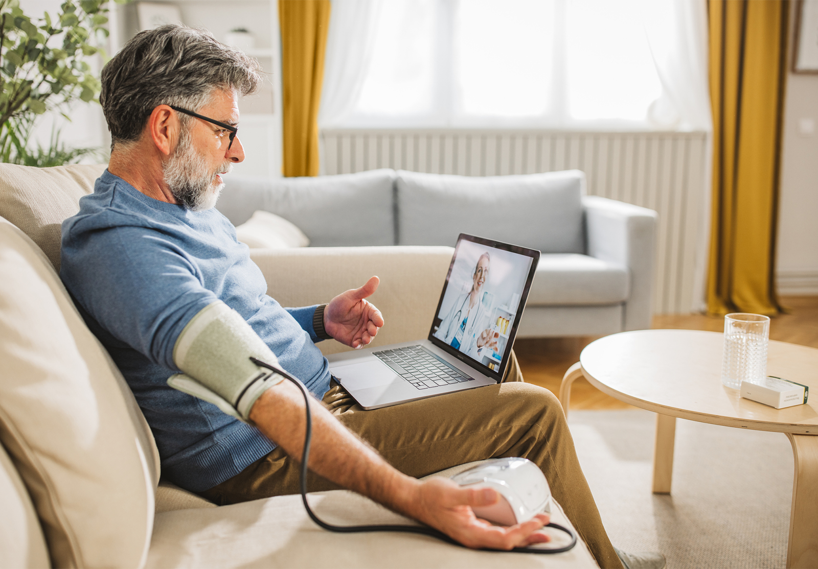 A man sits on a beige couch with a blood pressure cuff on his arm, speaking to a doctor via laptop in a sunlit living room, illustrating remote health monitoring and telemedicine.