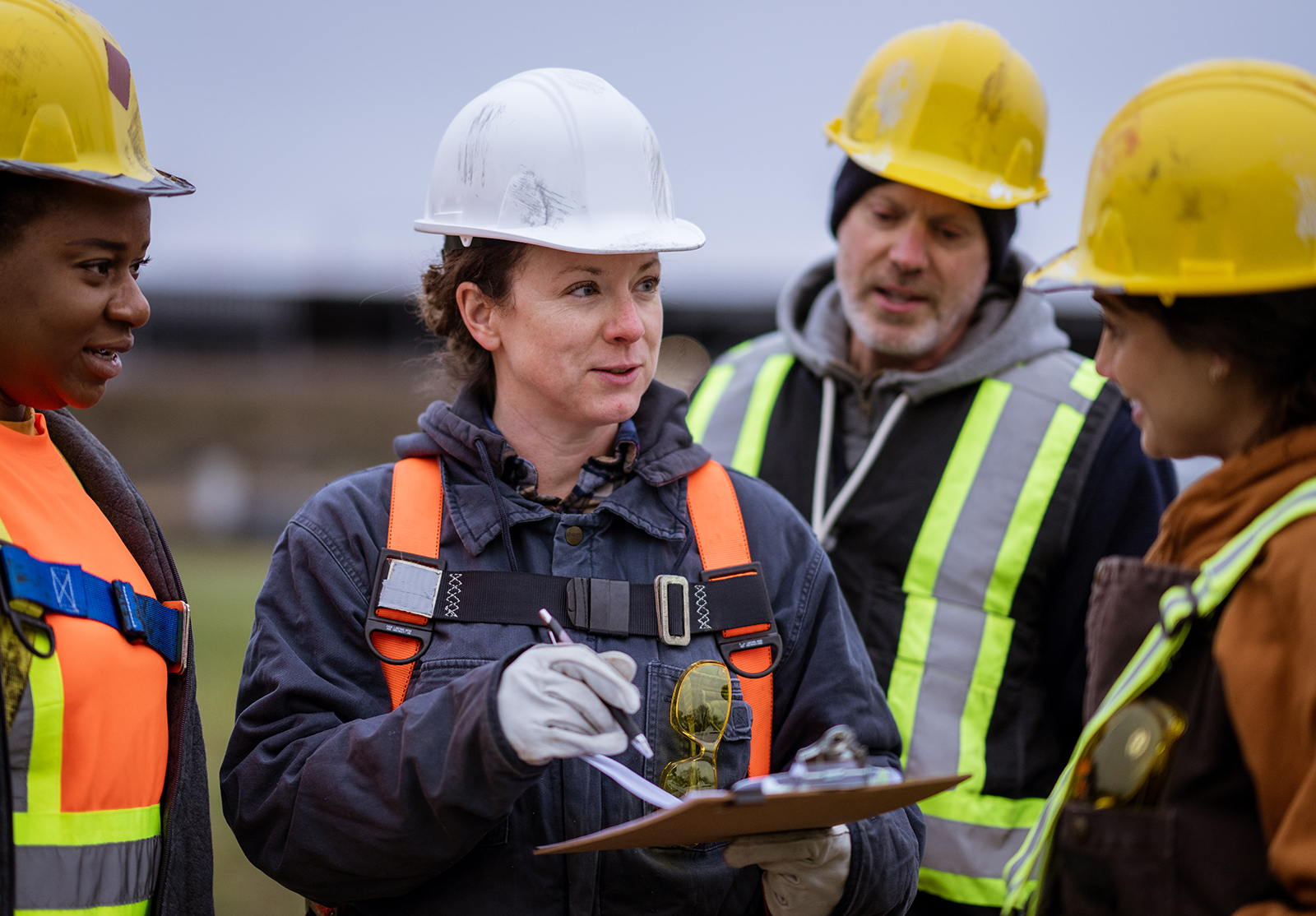 Construction workers in safety gear gather as one leads a site discussion with a clipboard.