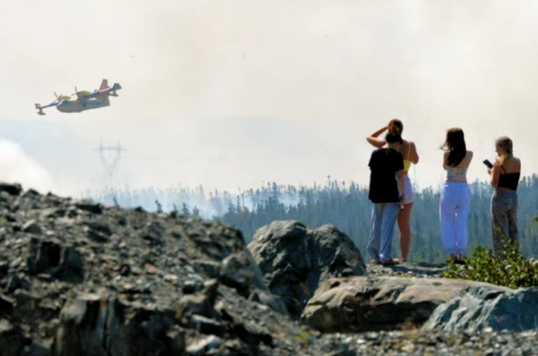At mid-day Tuesday August 12, 2025, at least three waterbombers, a birddog airplane and a helicopter were fighting the Paddy's Pond wildfire near St. John's, Mount Pearl and Paradise. Here, onlookers stand on a nearby hill watching the efforts. Photo by Keith Gosse/The Telegram (courtesy of The Canadian Press)