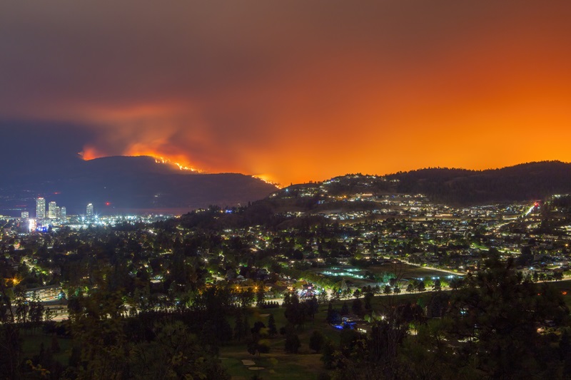 A wildfire burns on a mountainside near Kelowna illuminating the night sky with an orange glow while city lights shine below.