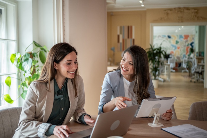 Two young businesswomen are working together on a project, using a laptop and a digital tablet, smiling and discussing in a modern office environment