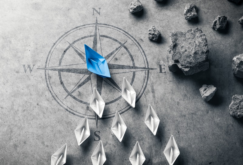 Blue Paper Boat With Compass Rose Leading A Fleet Of Small White Boats Away From Rocks On Grey Concrete - Leadership Concept