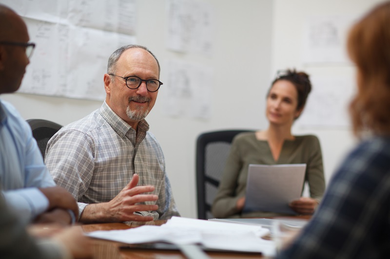 Senior man listens to coworker in meeting with work colleagues while sitting at table in design office