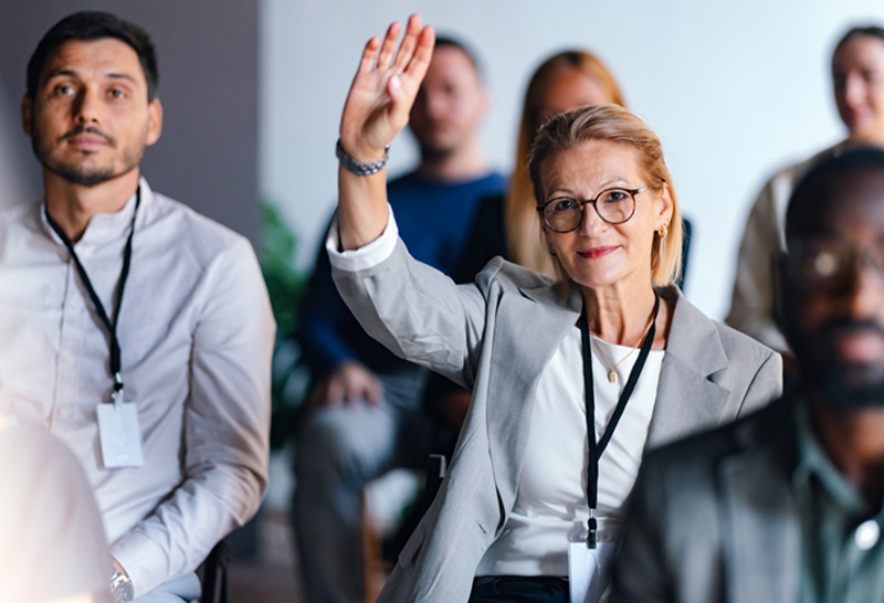 Focused businesswoman raises hand during a professional event, indicating engagement and participation. Diverse group of people in a coworking setting, highlighting teamwork and collaboration.
