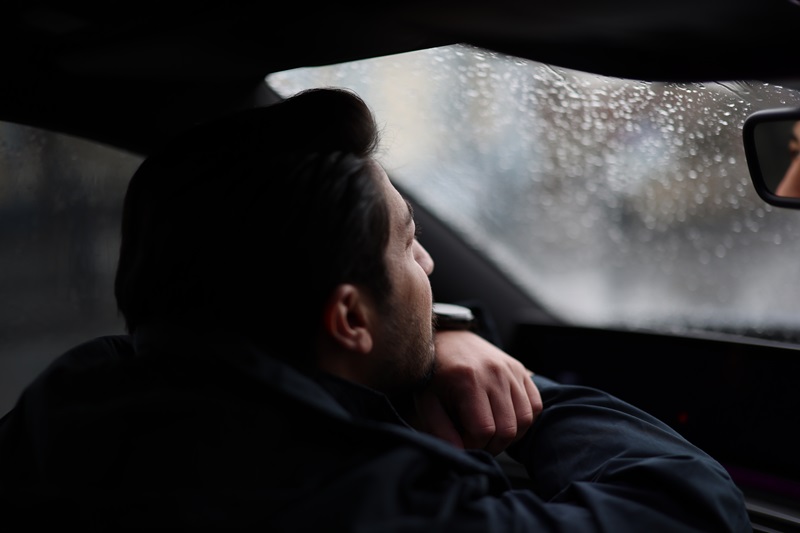 Man looking out a car window, resting his chin on his hand, observing rain falling. Feeling alone, contemplative and thoughtful